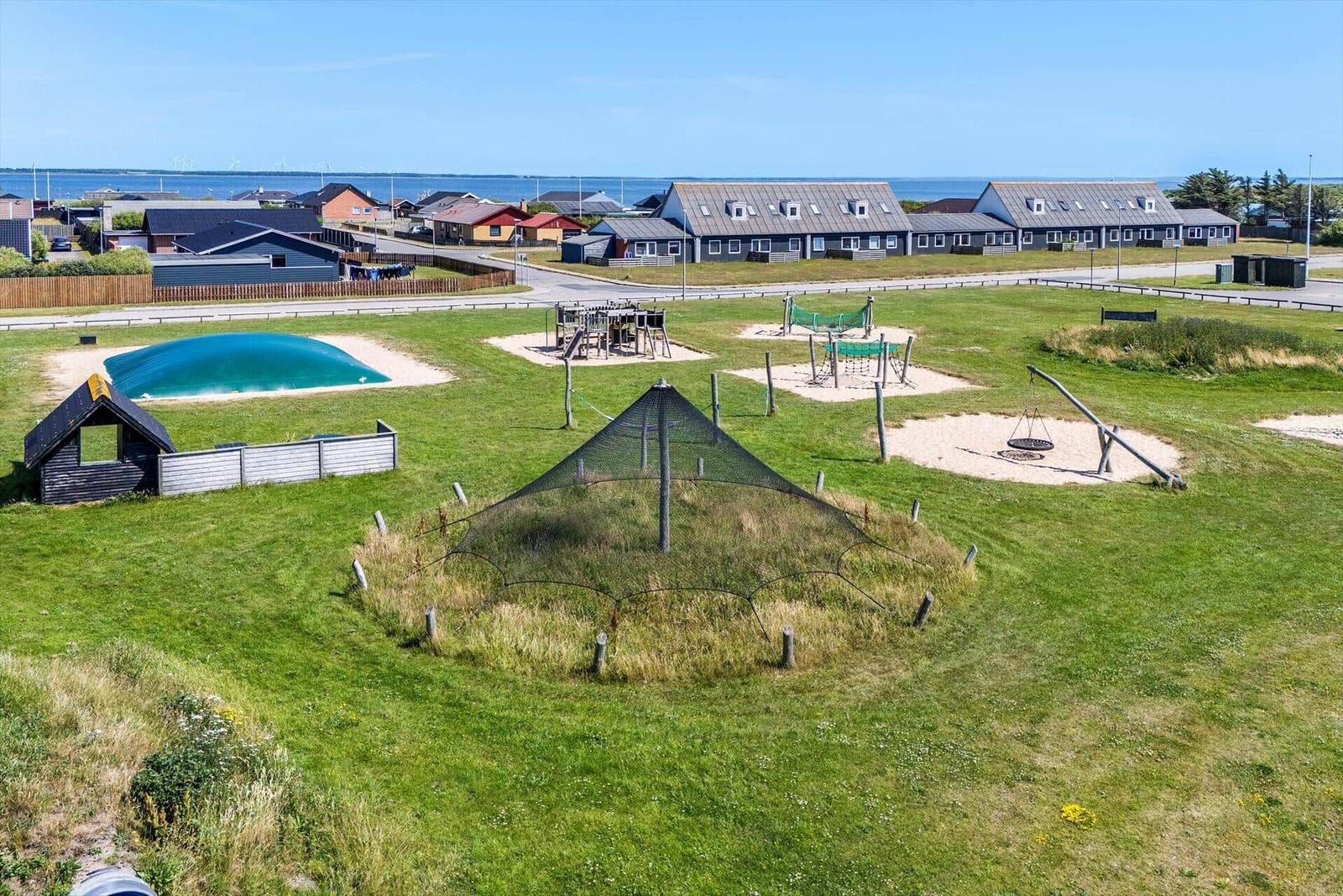 Grüner Spielplatz mit Sandkisten, Rutsche und Klettergerüsten. Hintergrund: Ferienhäuser und Meer.