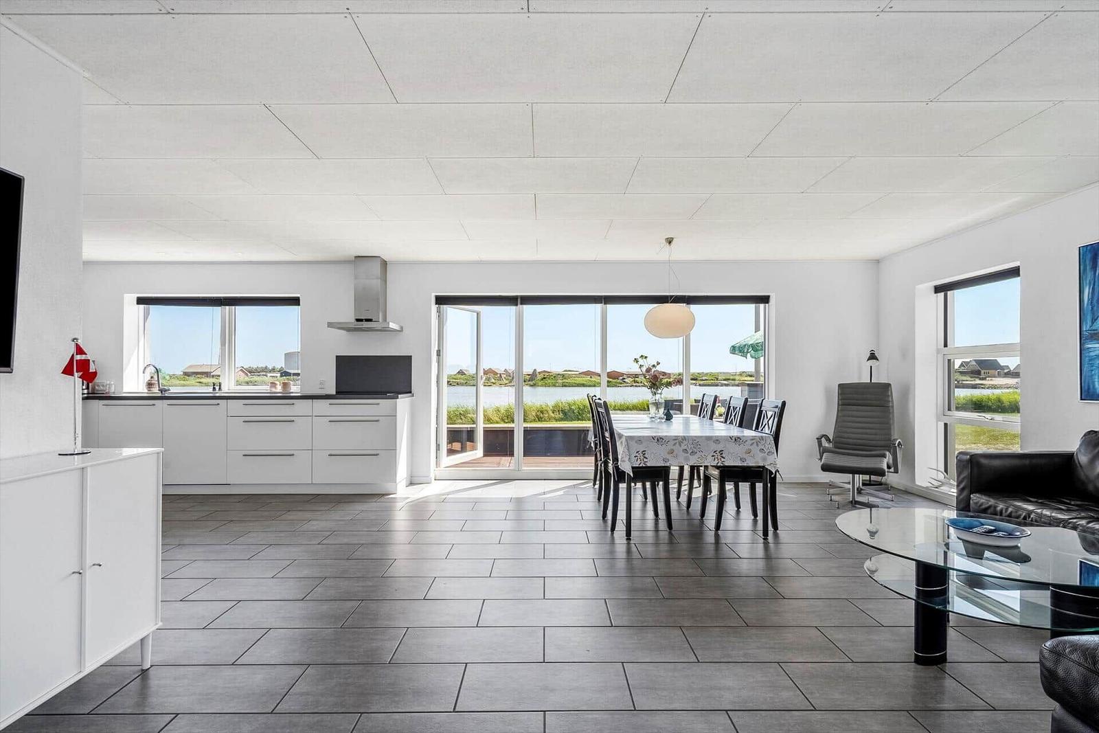 Offener Raum mit Küche, Esstisch und Sofa. Große Fenster mit Blick auf Wasser.