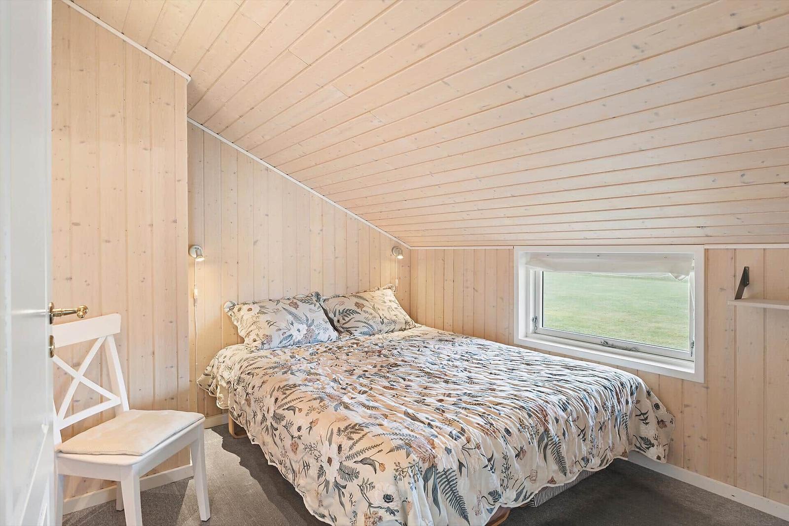 Schlafzimmer mit Bett, Fenster und Stuhl an Holzwand.