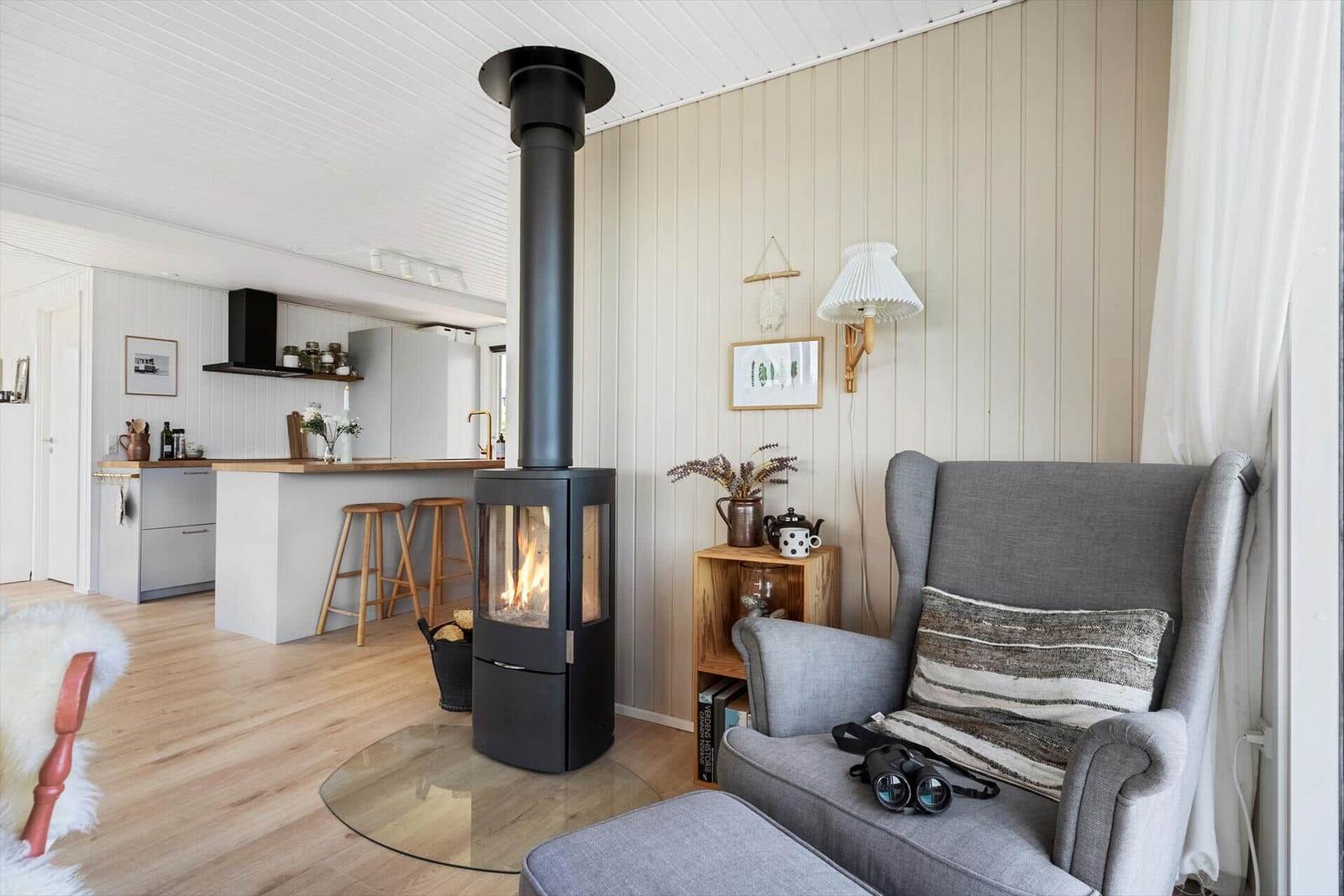 Wohnzimmer mit Kamin, grauer Sessel und Küchenbereich im Hintergrund.