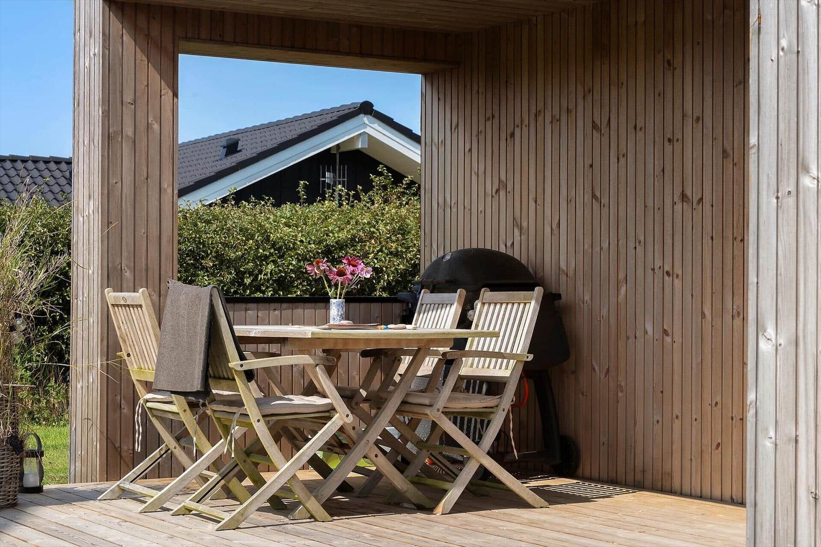 Terrasse mit Holztisch, vier Stühlen und Grill. Blick auf grüne Hecke und Haus.