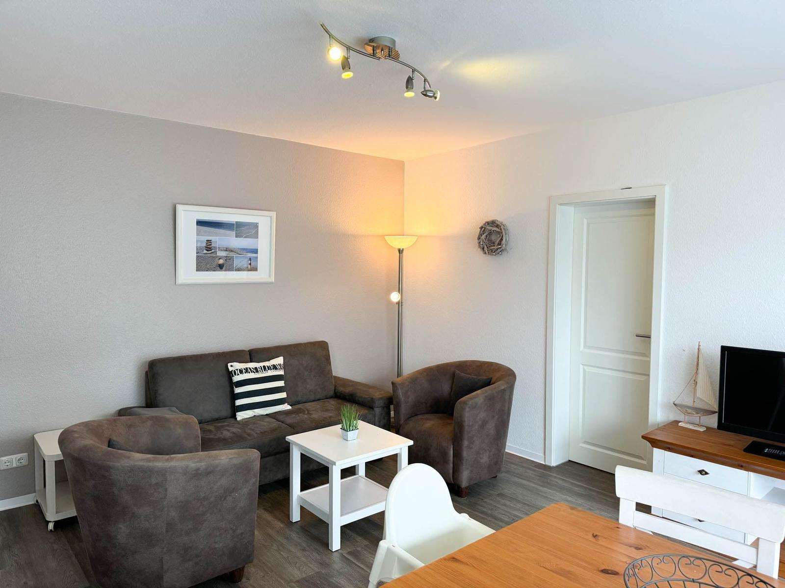 Wohnzimmer mit Sofa, Sesseln und Fernseher. Holztisch und Stehlampe sichtbar.