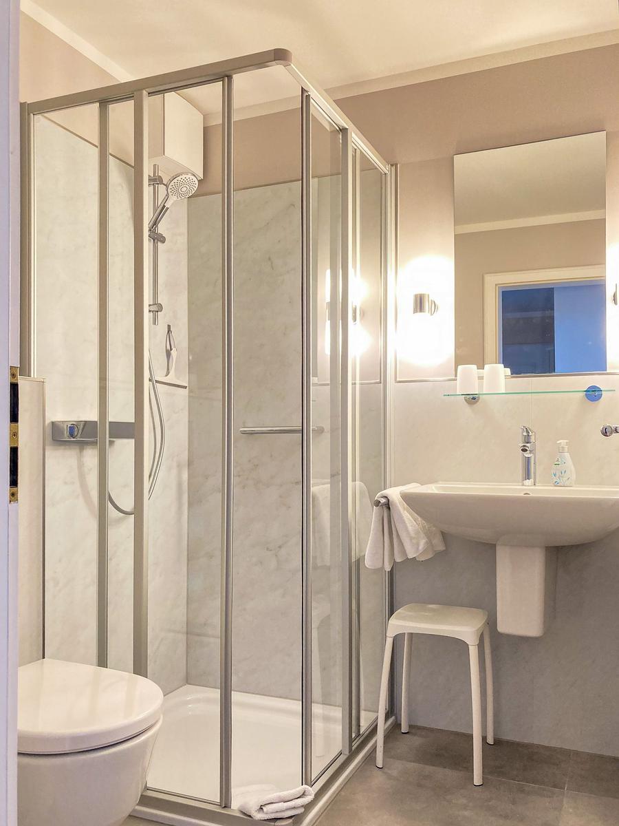Bad mit Dusche, Waschbecken und Toilette. Weiße Fliesen und Spiegel.