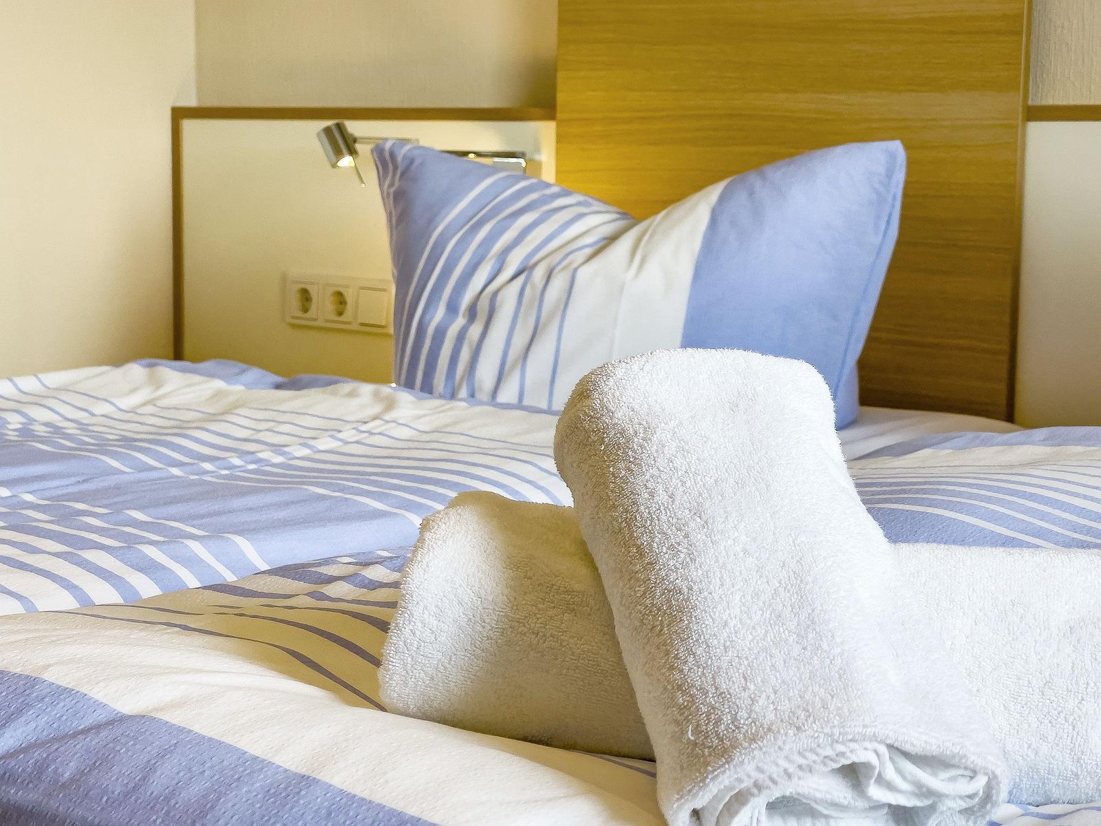Bettkopfteil aus Holz, Kissen und Bettwäsche mit blauen Streifen, gefaltete Handtücher auf dem Bett.