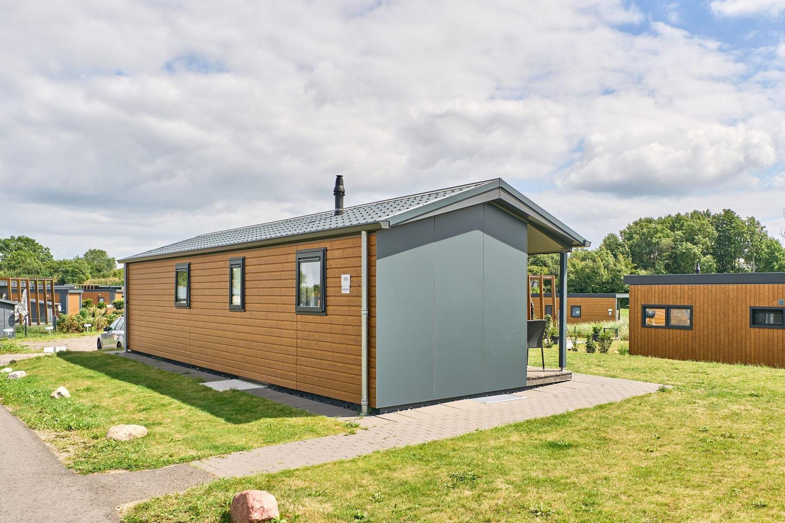 Moderne Holz-Hütte mit Terrasse und Garten. Dach mit Schornstein. Hintergrund mit Bäumen.