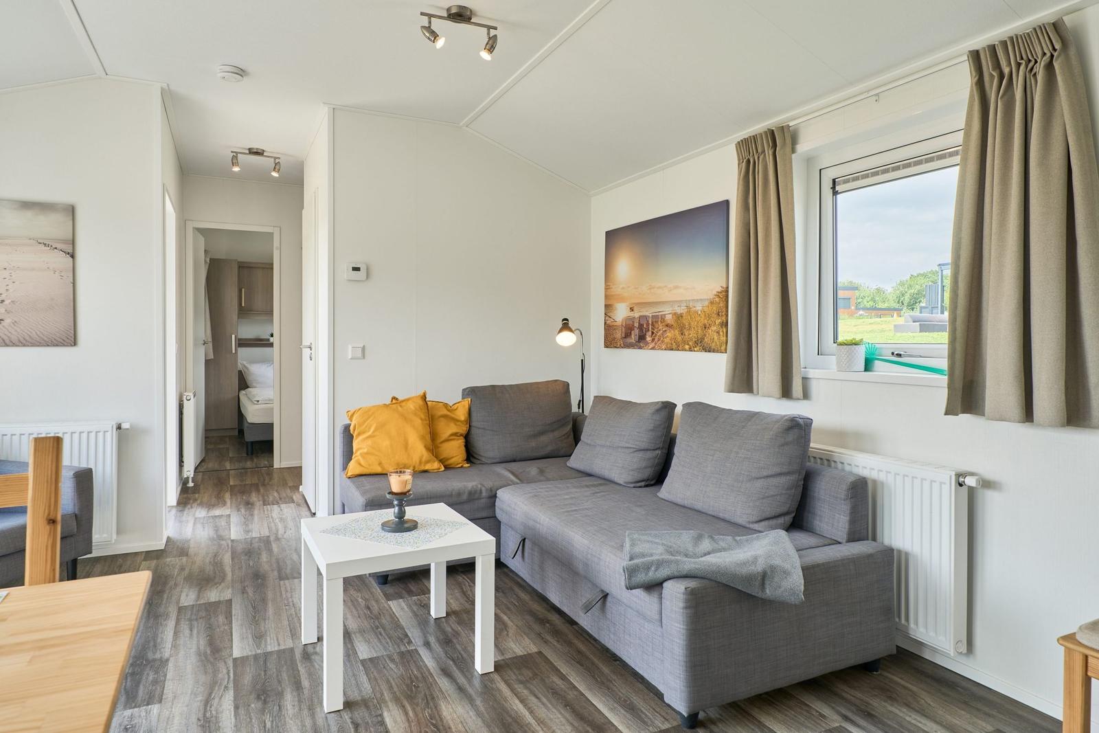 Wohnzimmer mit grauem Sofa, Tisch und Fenster mit Blick auf Grünflächen.