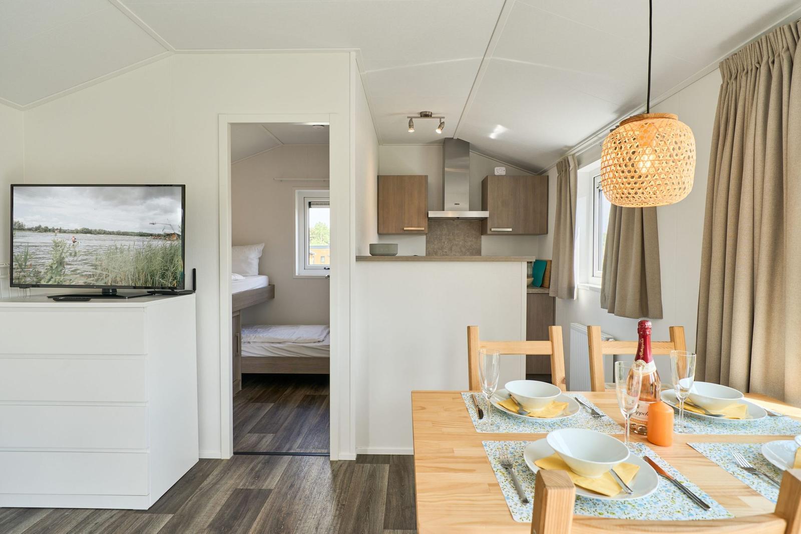 Küche, Essbereich und Schlafzimmer in einem Ferienhaus. Tisch gedeckt, TV eingeschaltet.