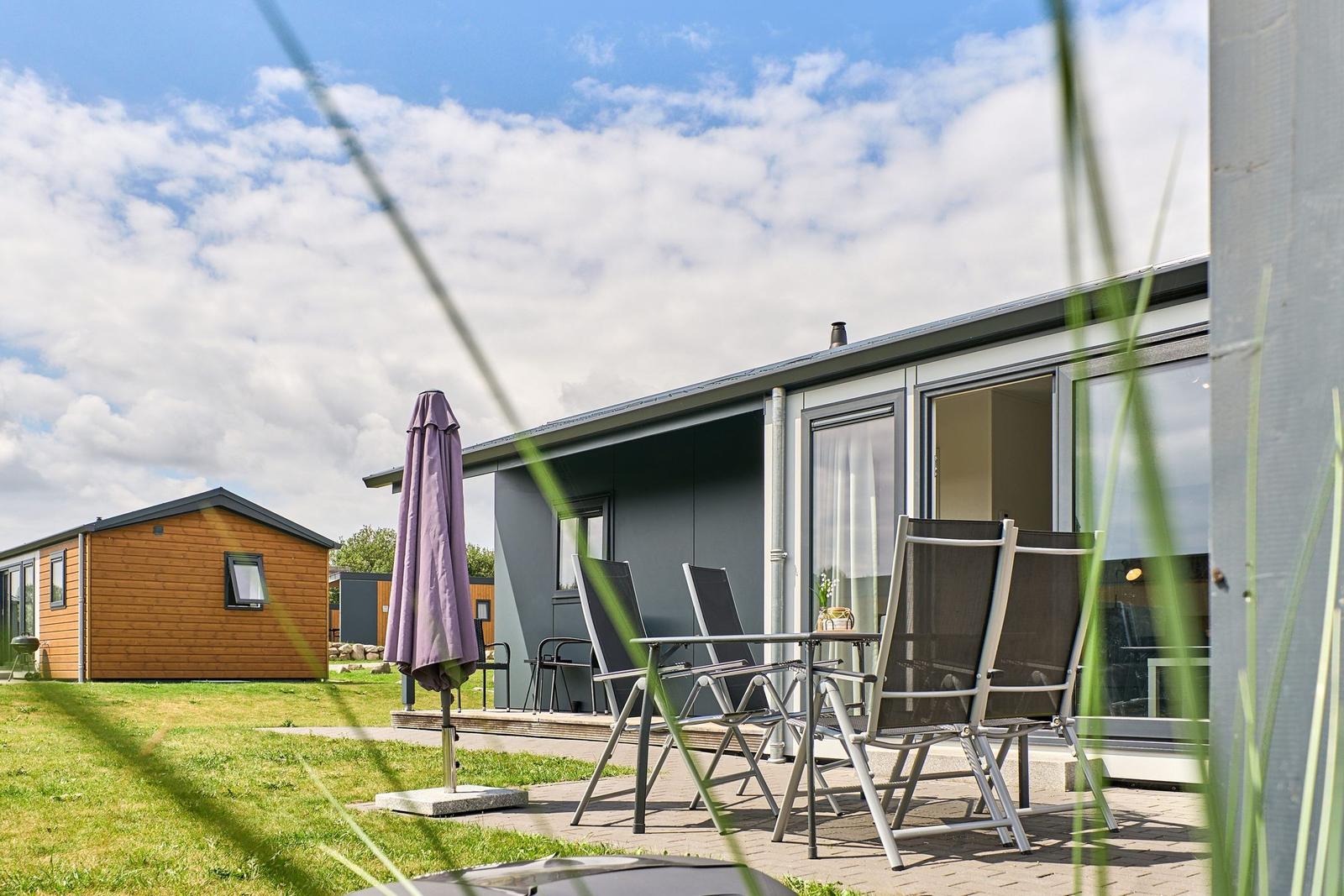 Terrasse mit Tisch, Stühlen und Sonnenschirm vor grauem Ferienhaus.