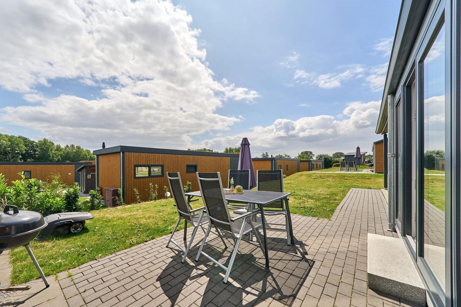 Terrasse mit Tisch, Stühlen und Grill. Hintergrund: moderne Holzhäuser und Grünfläche.