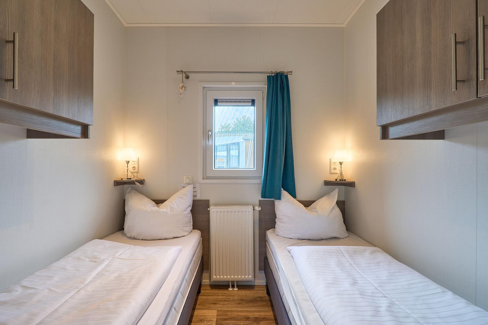 Zwei Einzelbetten mit weißen Bettwäsche, Fenster mit blauer Vorhang und Nachttische mit Lampen.