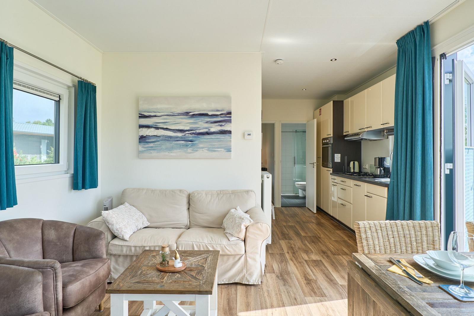 Wohnbereich mit Sofa, Couchtisch und Küchenbereich. Blaue Vorhänge und Holzboden.