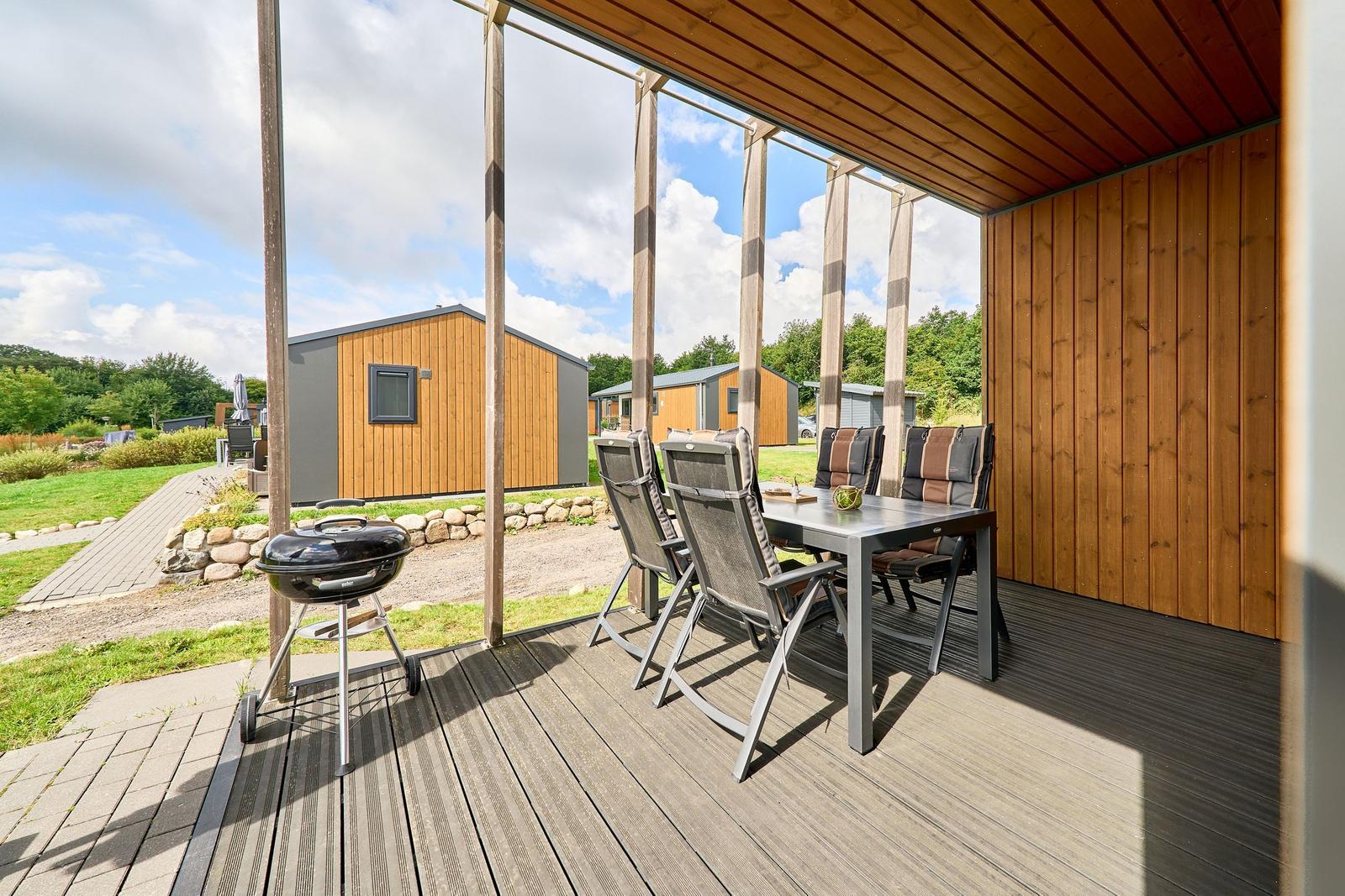 Terrasse mit Grill, Tisch und Stühlen. Hintergrund: Grasfläche und Holzhaus.