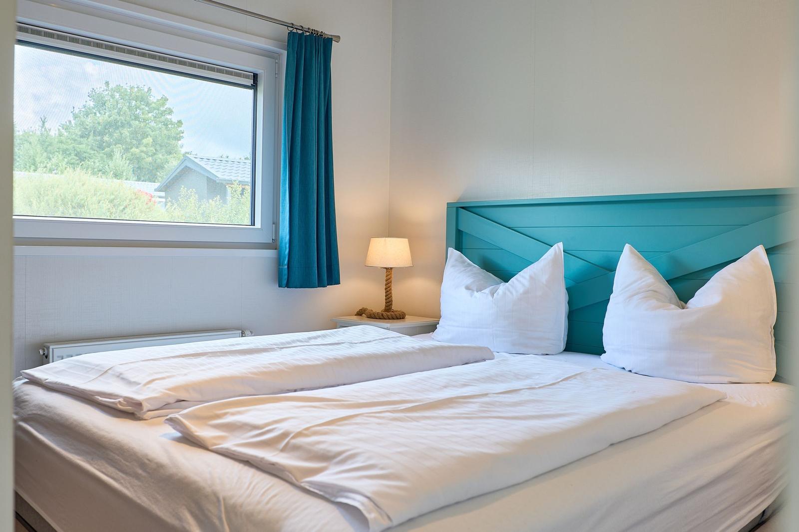 Schlafzimmer mit Doppelbett, blauem Kopfteil und Fenster mit Blick auf Grün.
