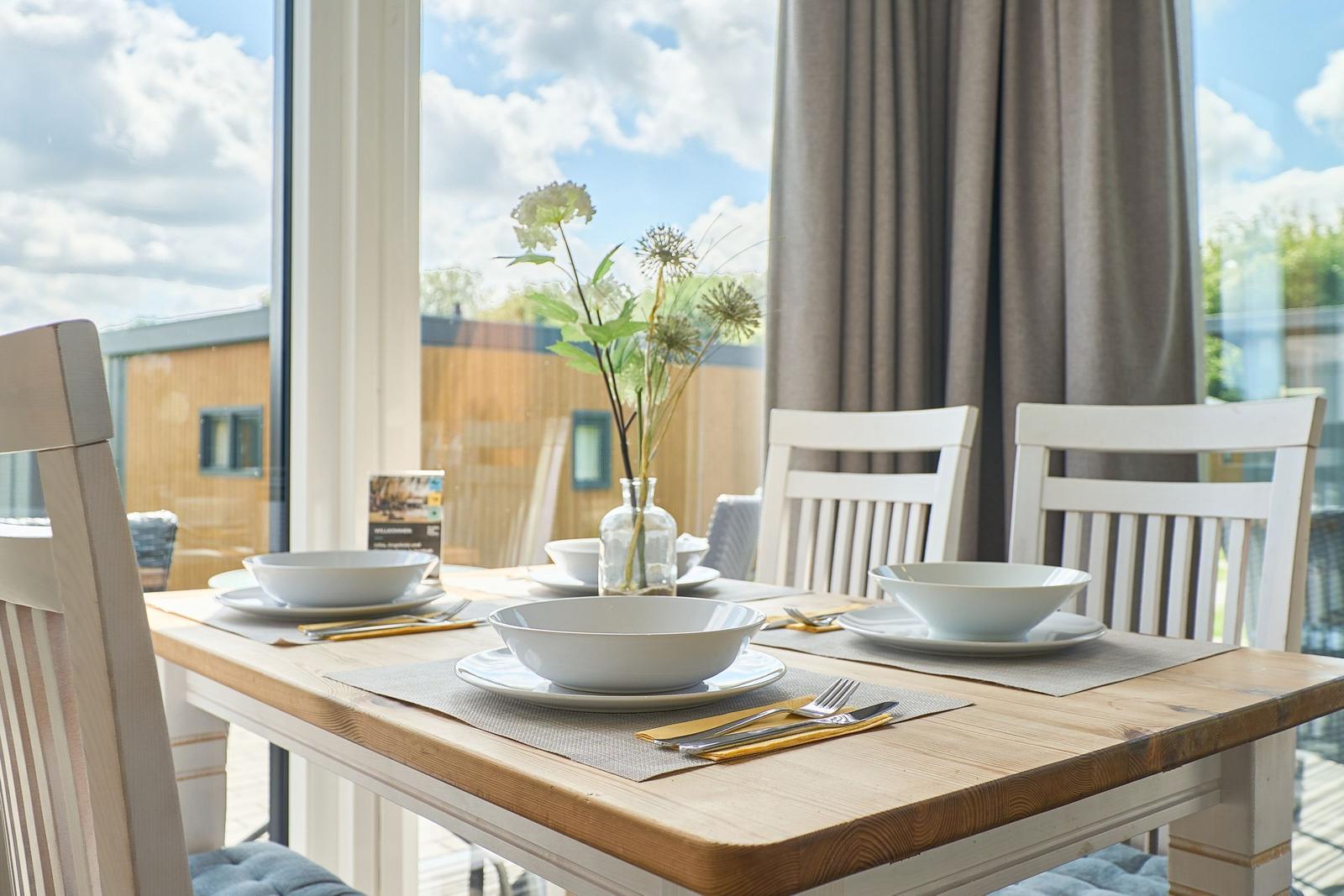 Esstisch mit Stühlen und Platz für vier Personen vor großen Fenstern.