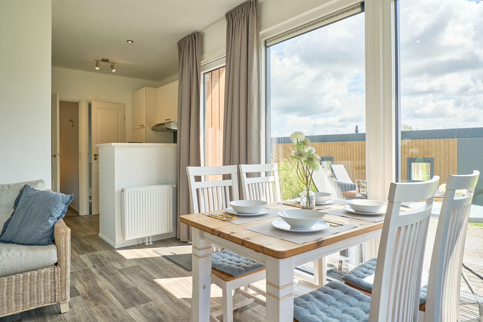 Essbereich mit Tisch und Stühlen vor großem Fenster. Küche und Wohnbereich sichtbar.