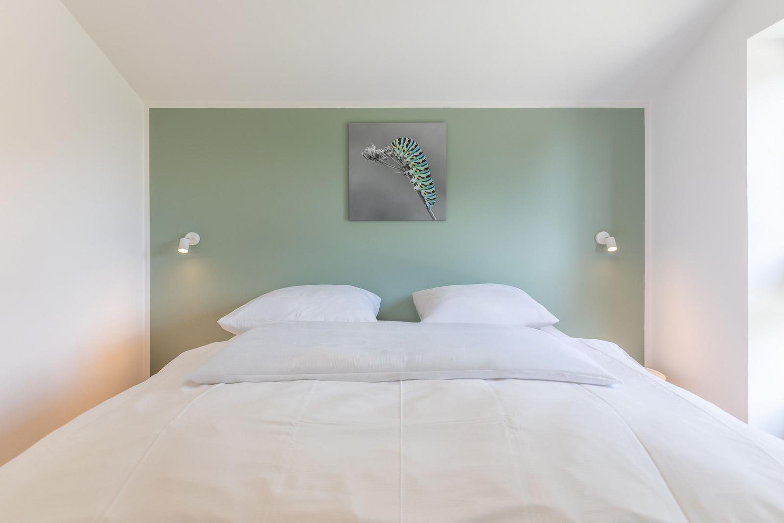 Doppelbett mit weißer Bettwäsche, grüner Wand und Wandbild mit Schmetterlingsraupe.
