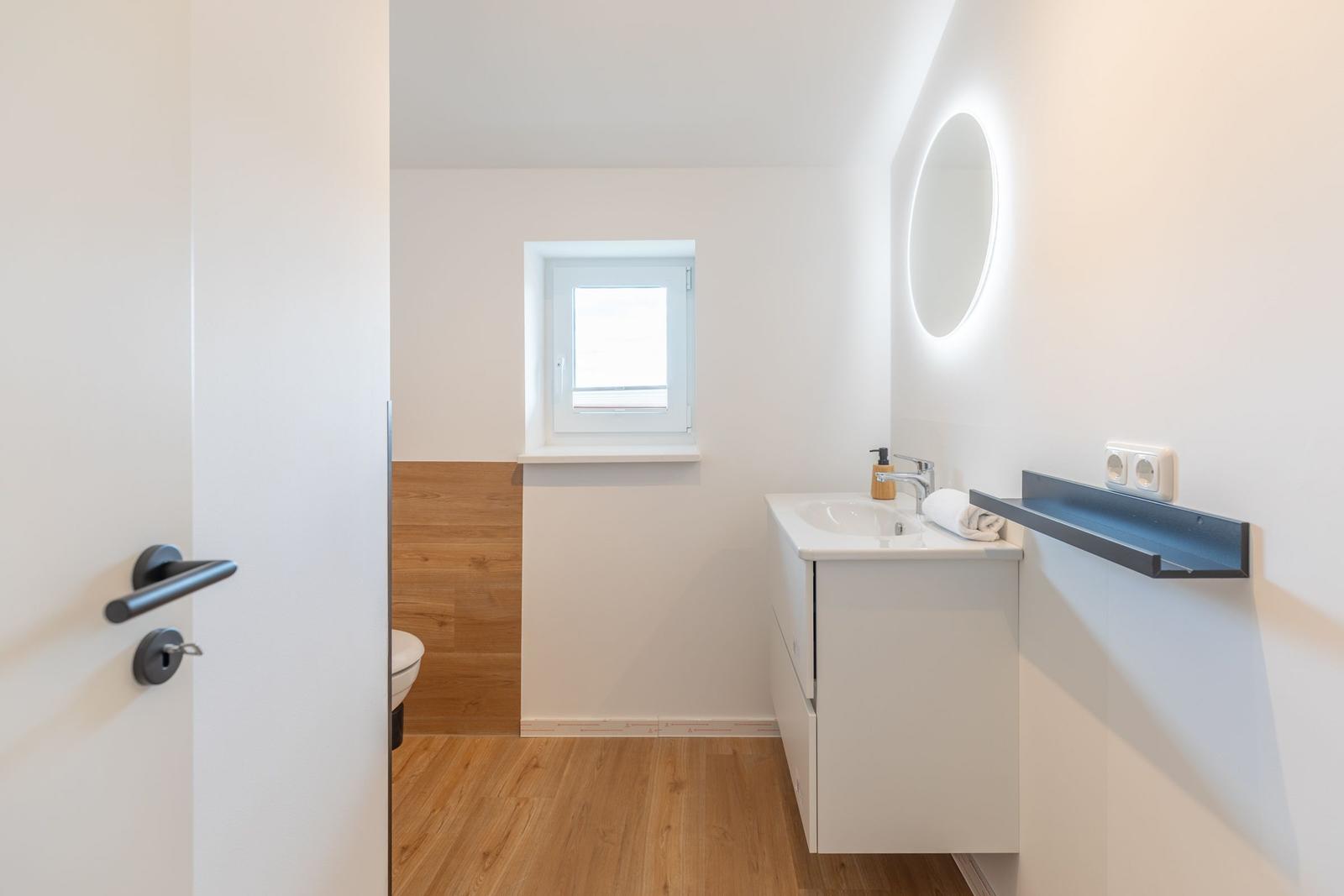 Badezimmer mit Waschbecken, Spiegel und Toilette. Holzboden und Fenster.