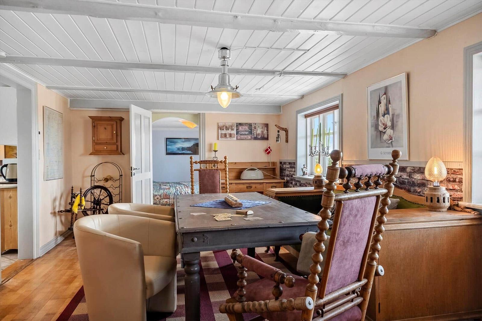 Dining room with wooden table, chairs, and window. Ceiling with beams. Wall with painting.
