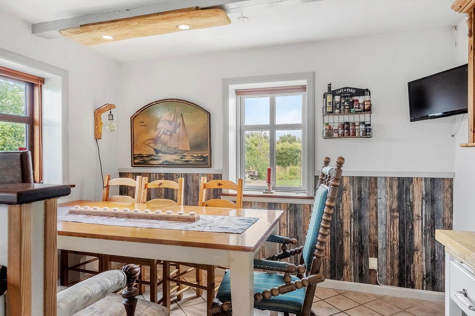 Dining room with wooden table, chairs, and wall painting. Windows with view of greenery.