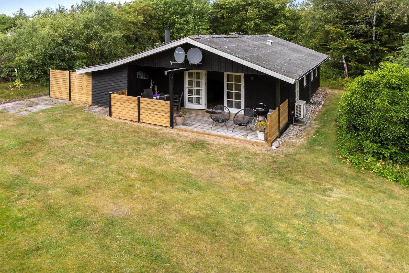 Black wooden house with terrace, grill, and garden. Surrounded by trees.
