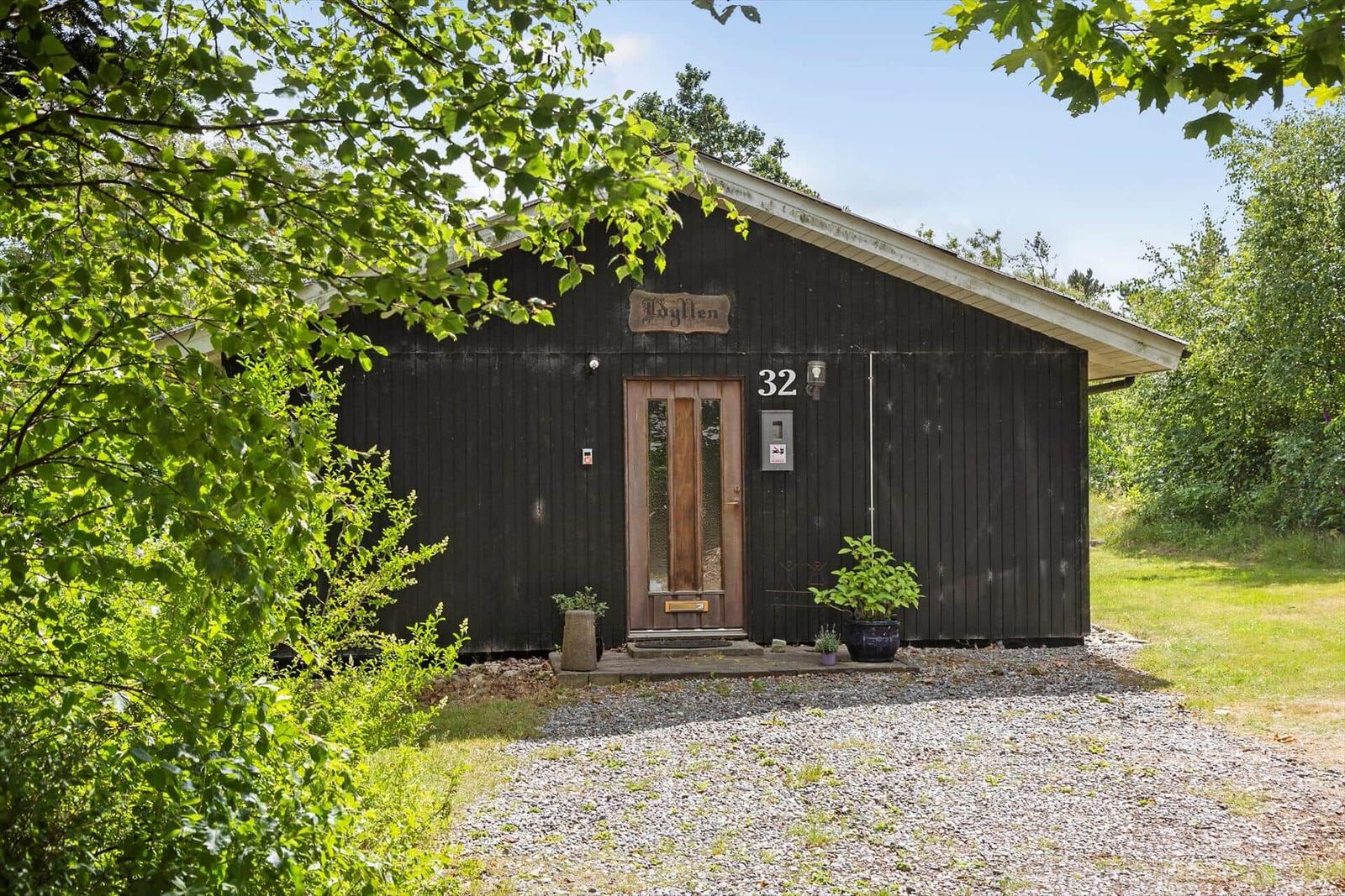 Black wooden house with entrance door and number 32. Surrounded by trees and grass.