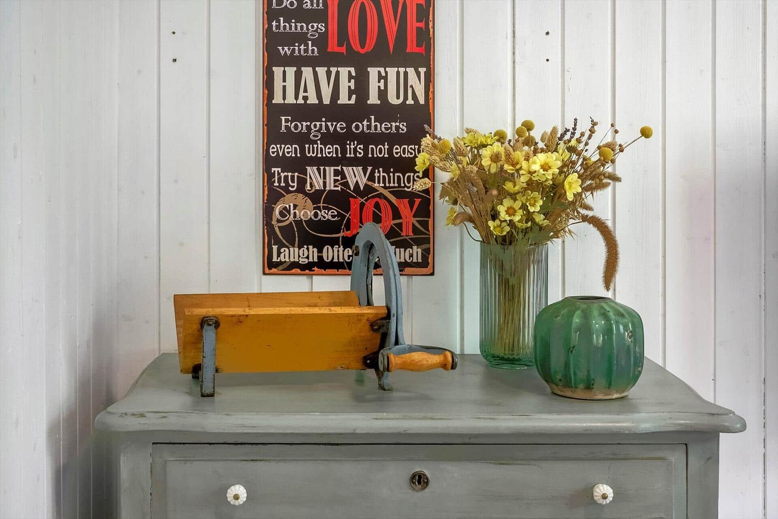 A gray dresser with flowers, vase, and wooden object against white wall.