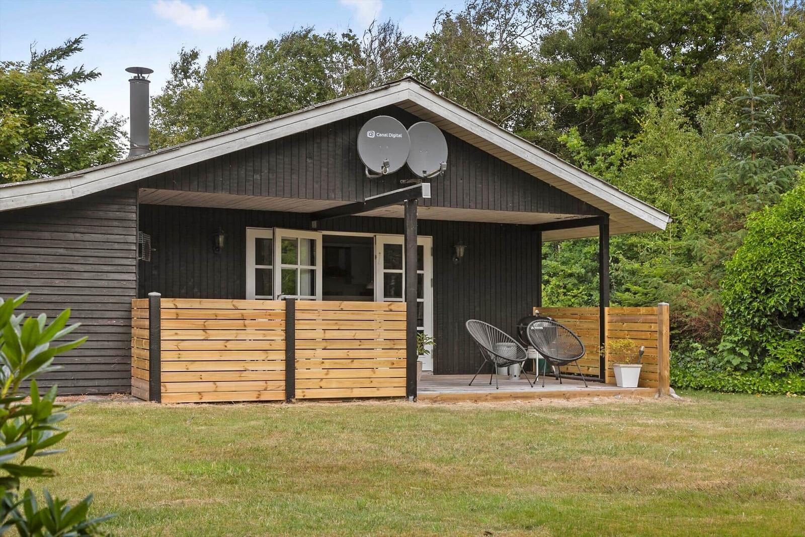 Wooden porch with two chairs and grill in front of a dark wooden house with satellite dishes.