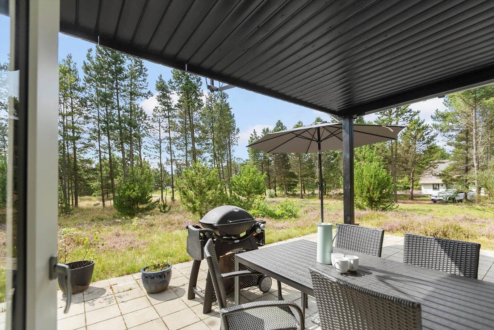 Terrasse mit Tisch, Stühlen, Sonnenschirm und Grill. Blick in den Wald.
