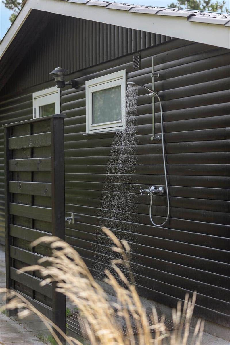 Außen-Dusche an dunklem Holzhaus mit Fenster und Pflanzen im Vordergrund.