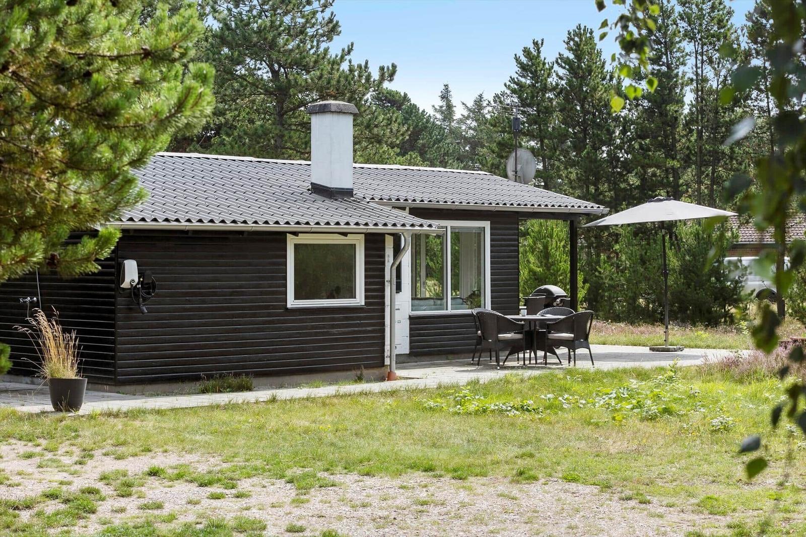 Ein dunkelholzverkleidetes Haus mit Terrasse, Grill und Sonnenschirm im Wald.