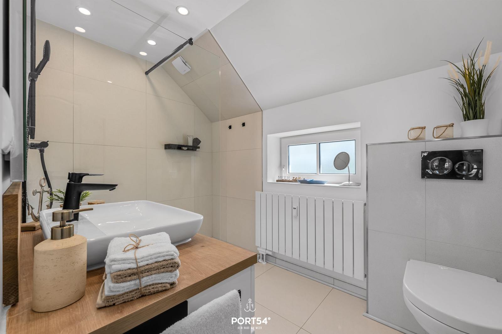 Badezimmer mit Dusche, Waschbecken und Toilette. Fenster mit Pflanze und Heizkörper.