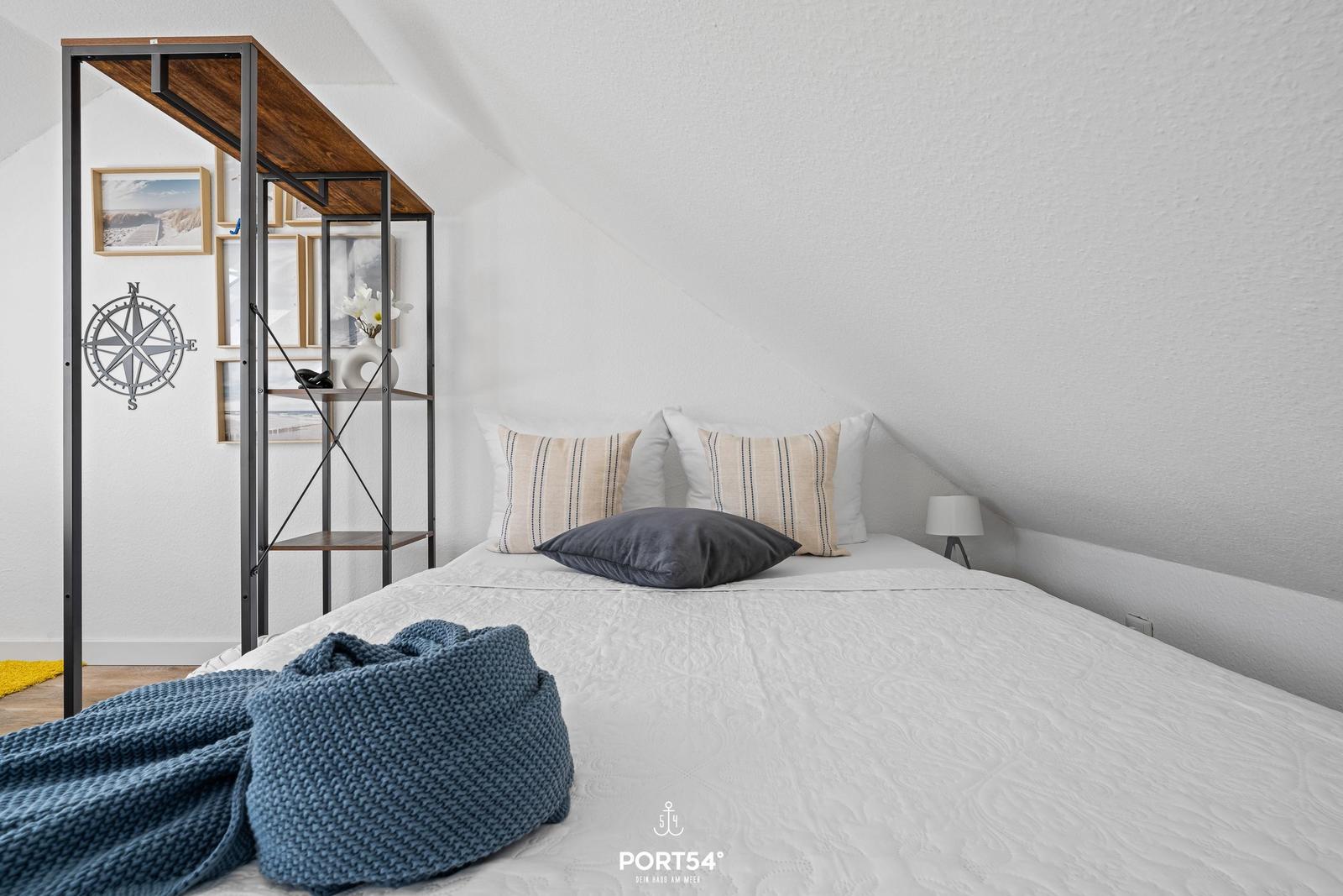 Schlafzimmer mit Bett, Kissen und blauer Decke. Wandregal mit Bildern und Kompass.