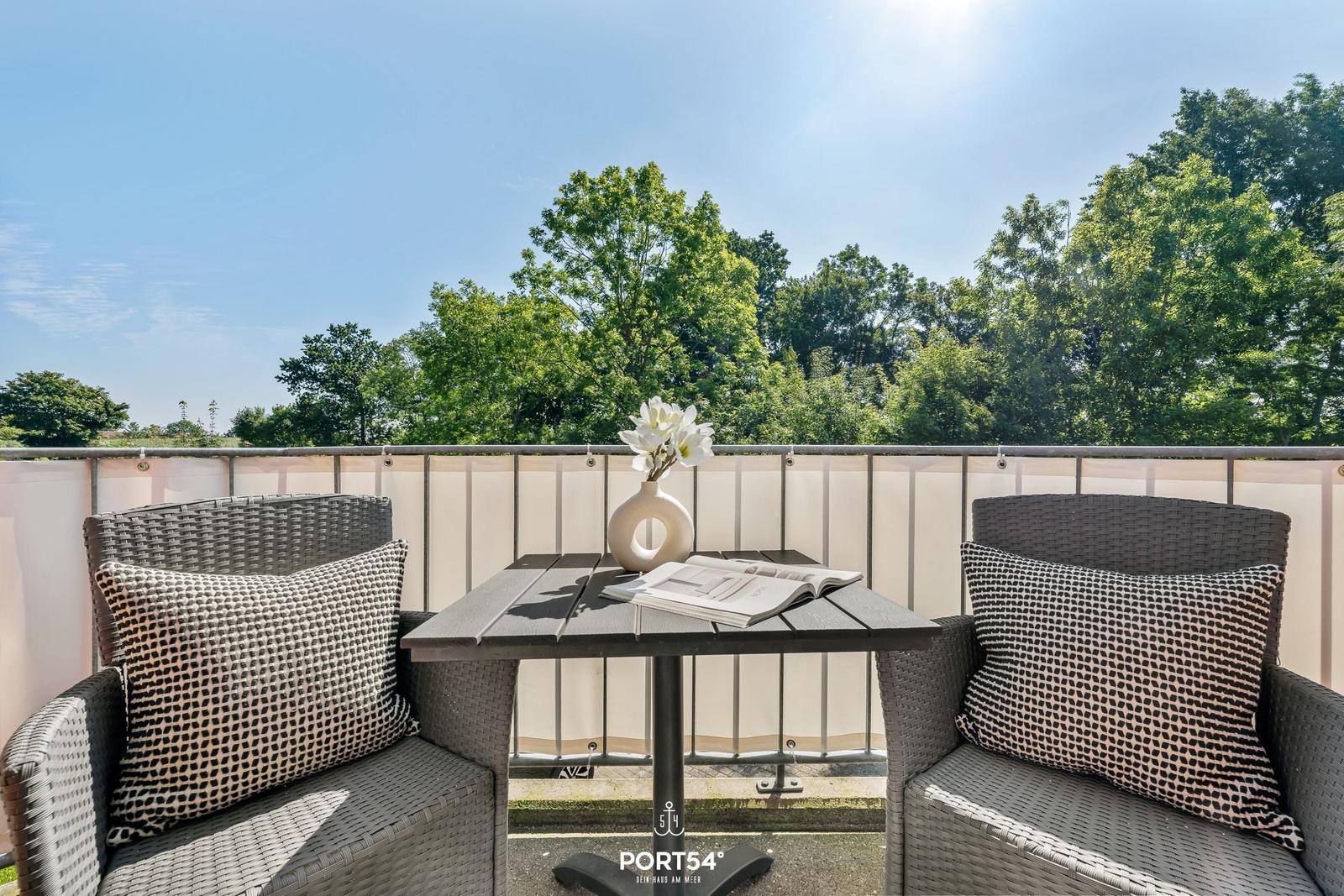 Terrasse mit zwei Stühlen, Tisch und Blumen. Blick auf grüne Bäume.