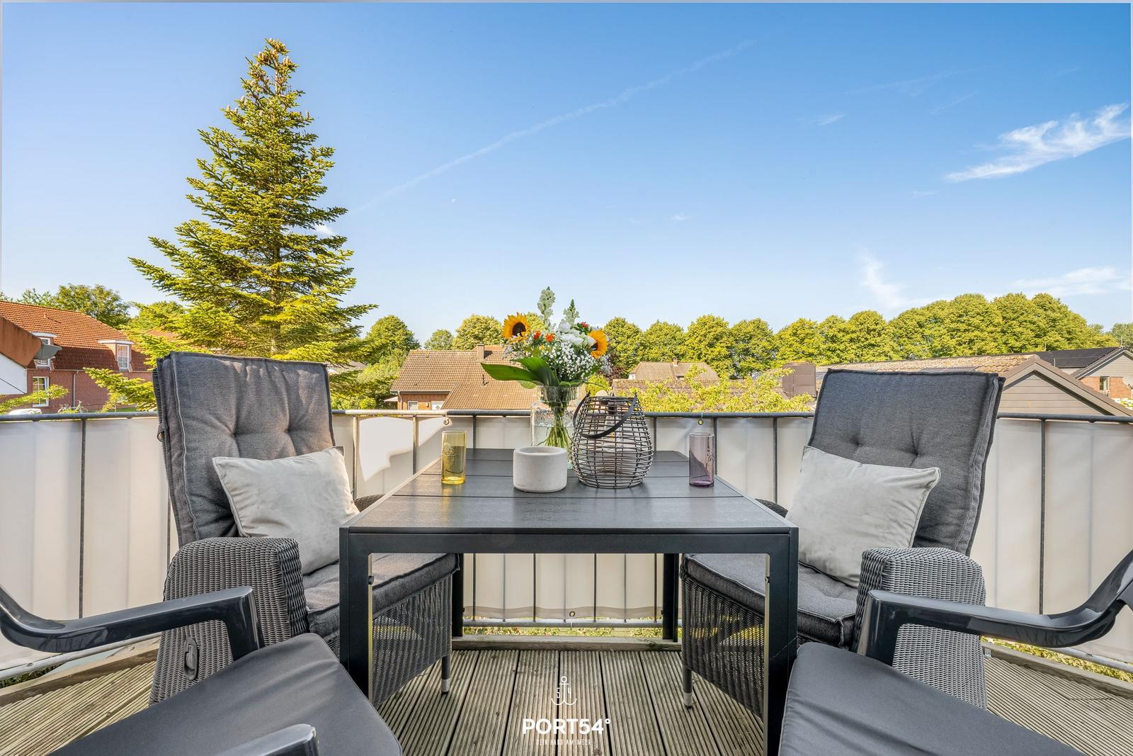 Terrasse mit zwei Sesseln, Tisch und Blumenarrangement. Blick auf Bäume und Häuser.