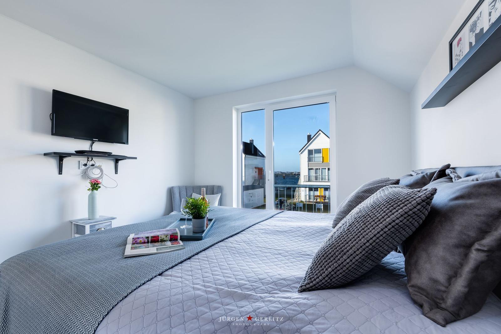 Schlafzimmer mit Blick aufs Wasser und Balkon. Bett, TV und kleiner Tisch.