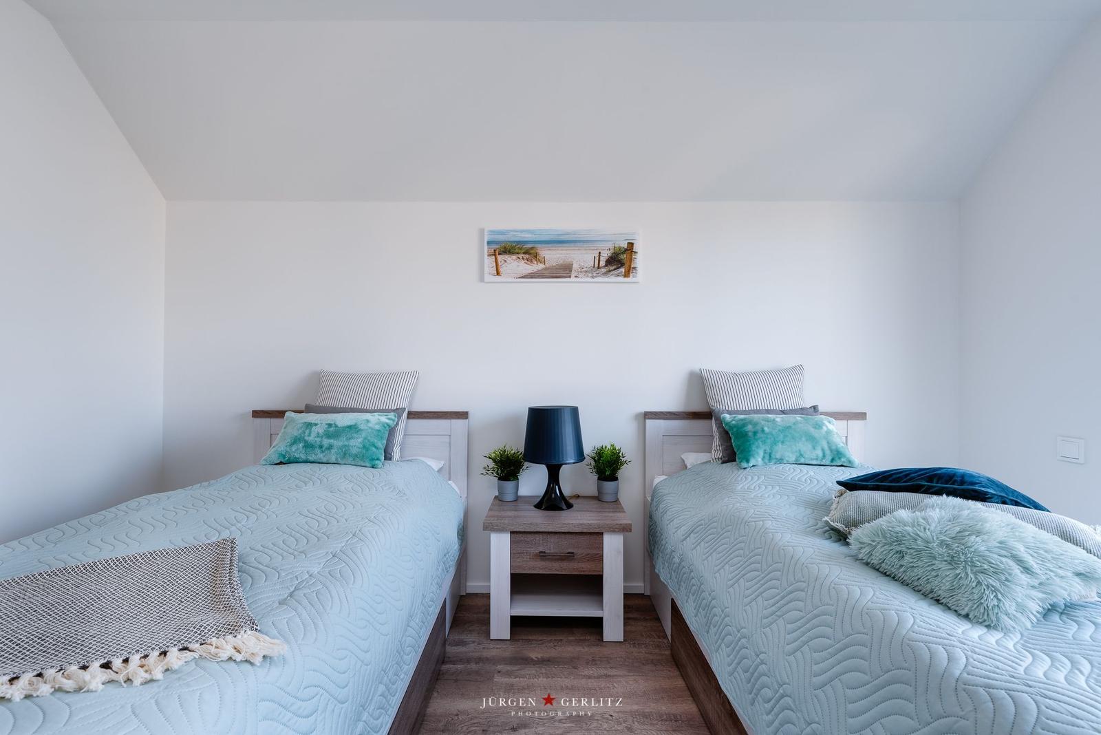 Zwei Betten mit hellblauen Bettwäsche und Kissen. Zwischen ihnen steht ein Nachttisch mit Lampe und Pflanzen.