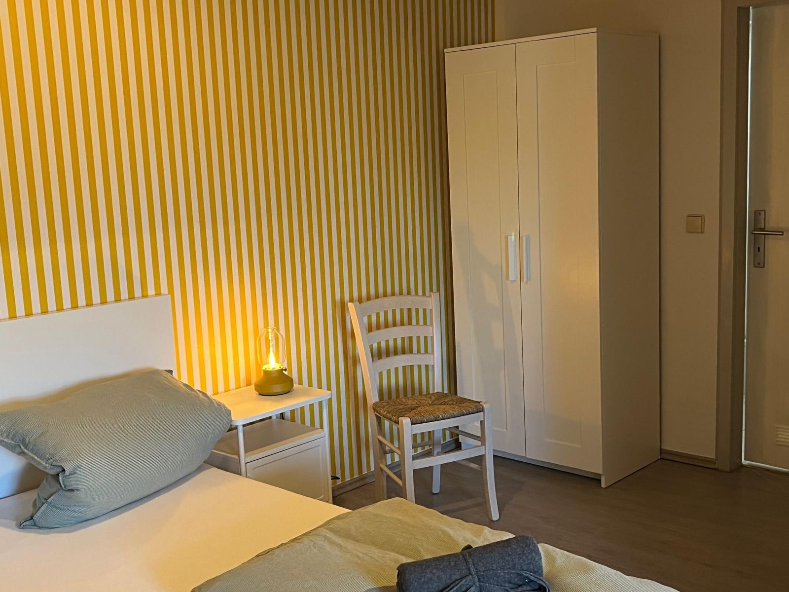 Schlafzimmer mit gelb-weiß gestreifter Wand, Bett, Nachttisch, Stuhl und Kleiderschrank.