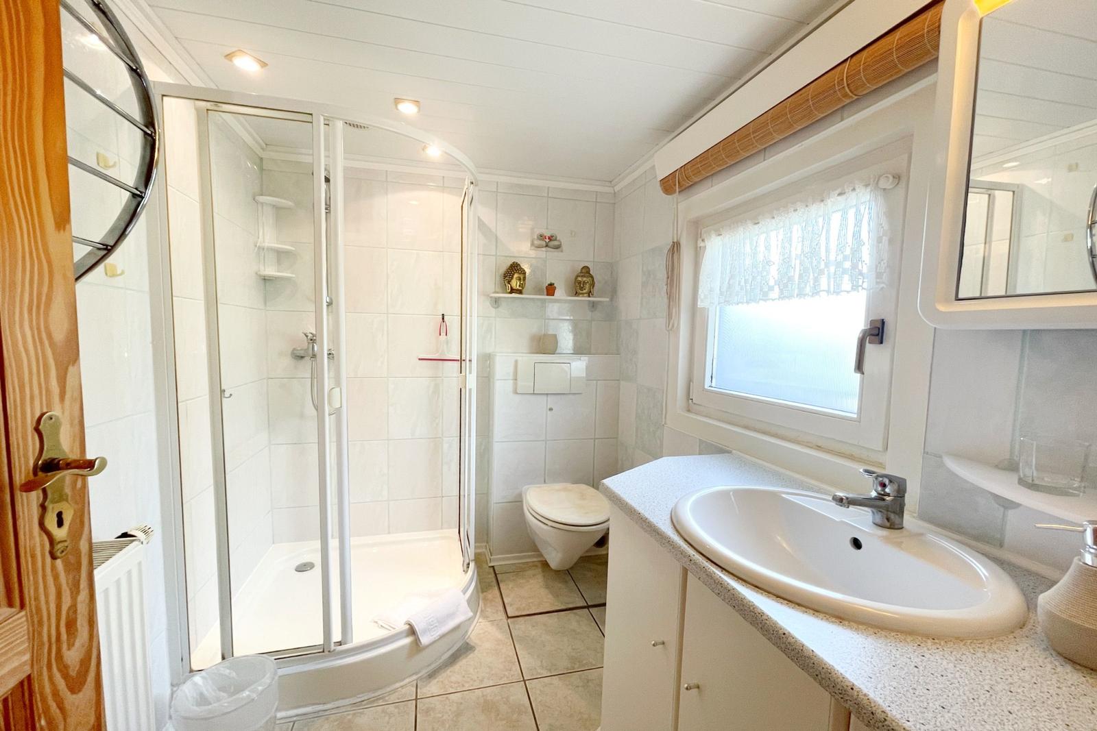 Badezimmer mit Dusche, Waschbecken und Toilette. Fenster mit Vorhang.