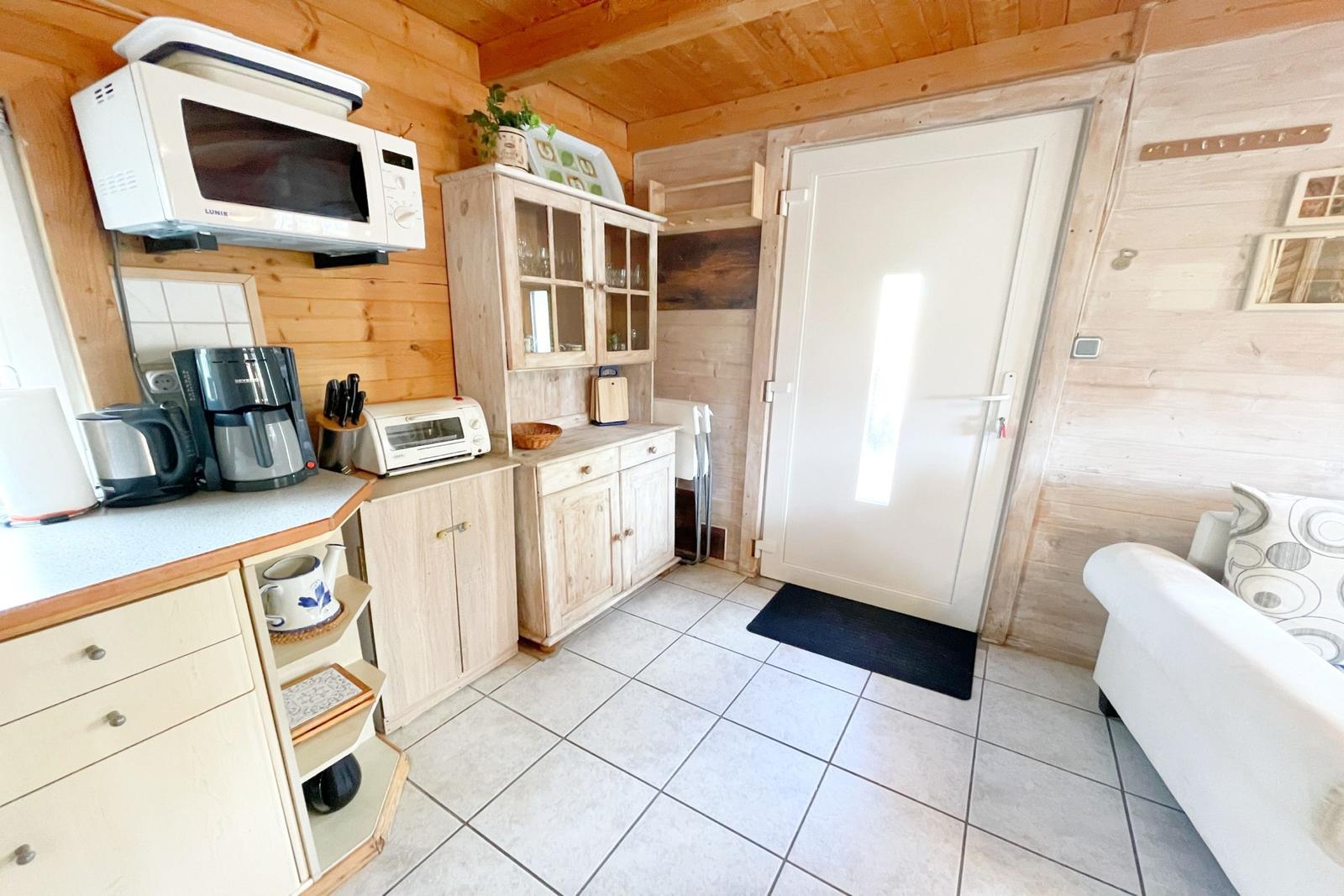 Küche mit Mikrowelle, Kaffeemaschine und Küchenzeile. Tür und Sofa sind sichtbar.