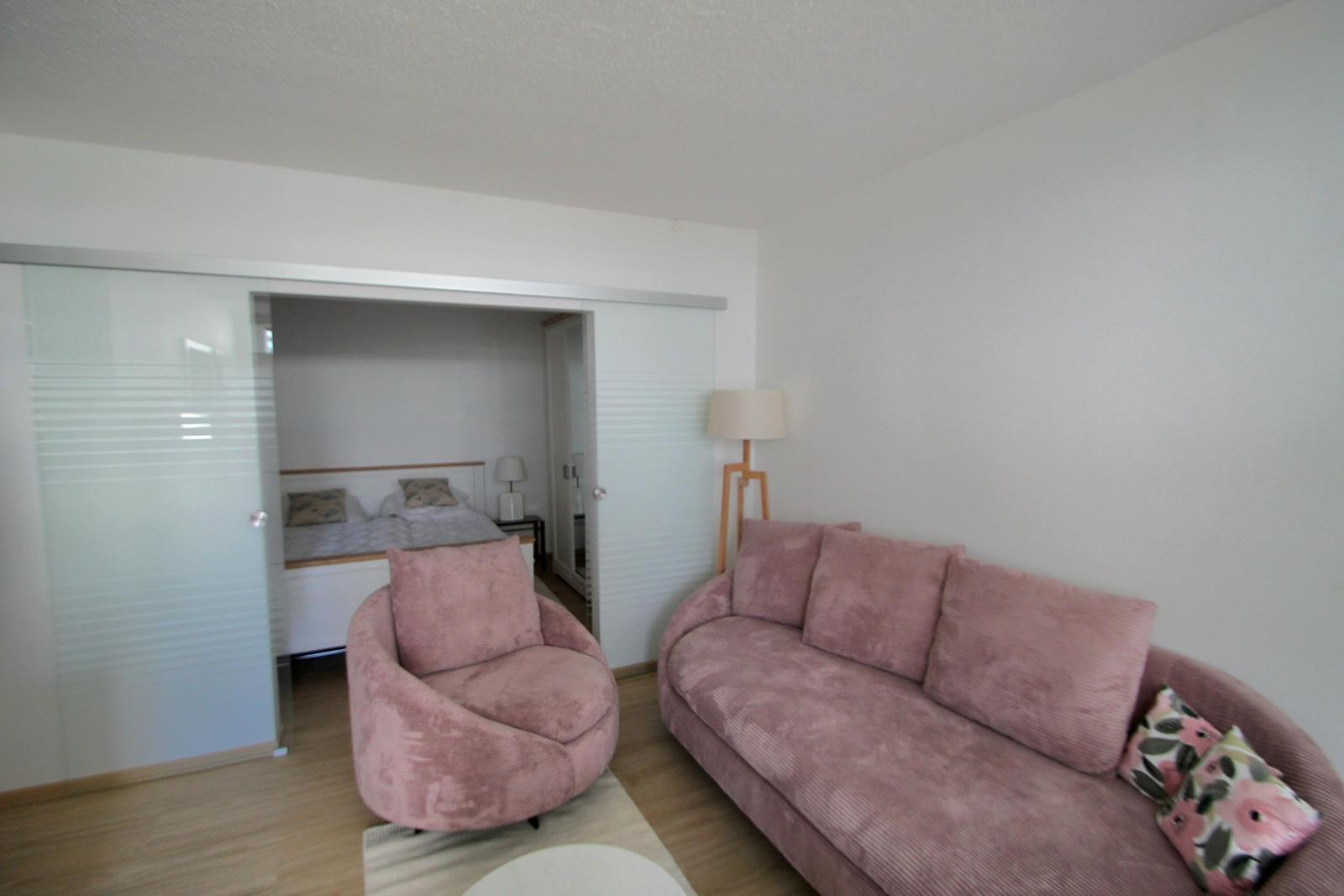 Wohnzimmer mit rosa Sofa und Sessel. Durchgehendes Schlafzimmer mit Bett sichtbar.