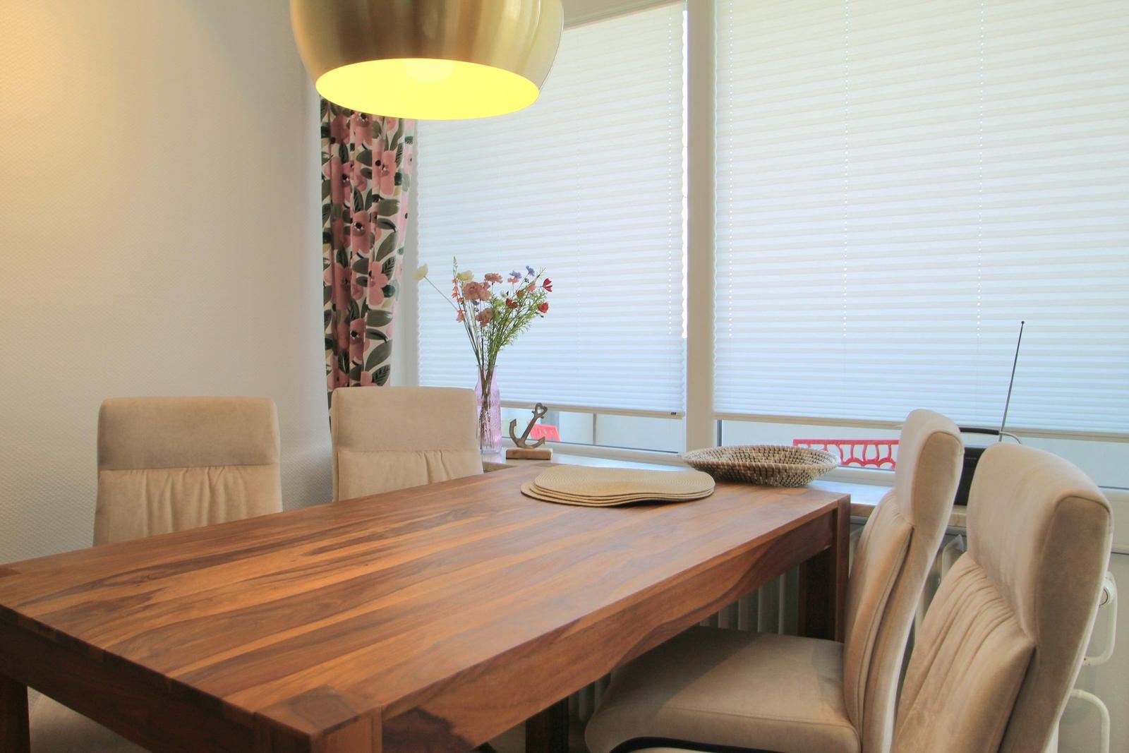 Esstisch mit Stühlen vor Fenster mit Jalousien und Blumenvase.