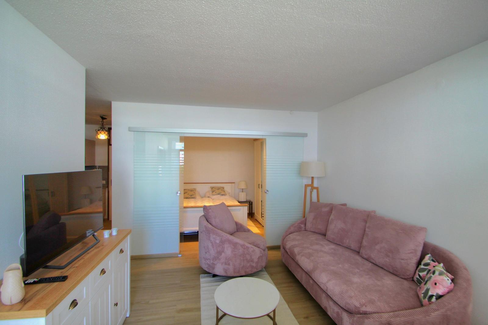 Wohnbereich mit Sofa, Sessel und TV. Schlafzimmer dahinter durch Glaswand abgetrennt.