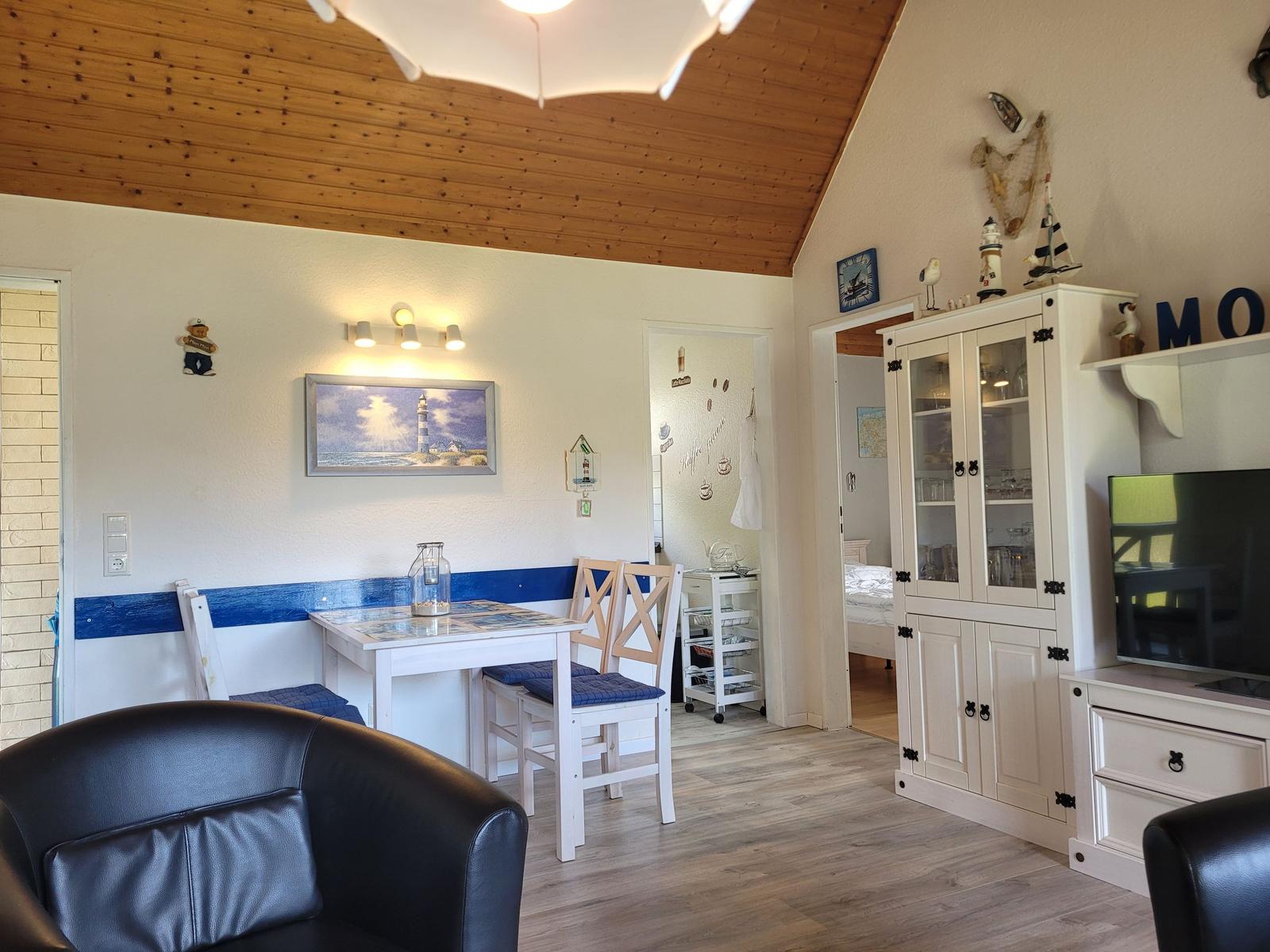 Wohnzimmer mit Esstisch, TV und Küchenzeile. Holzdecke und weiße Möbel.