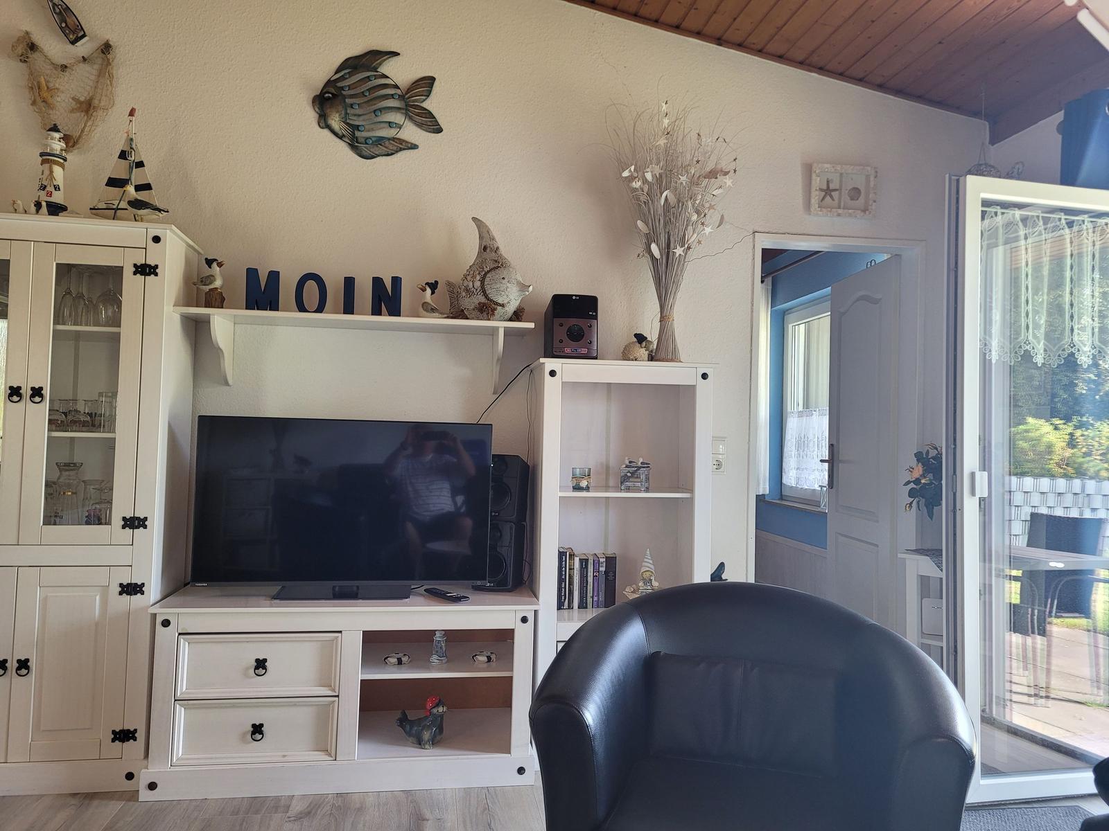 Wohnzimmer mit Fernseher, Schrank und Sessel. Tür führt auf Balkon.