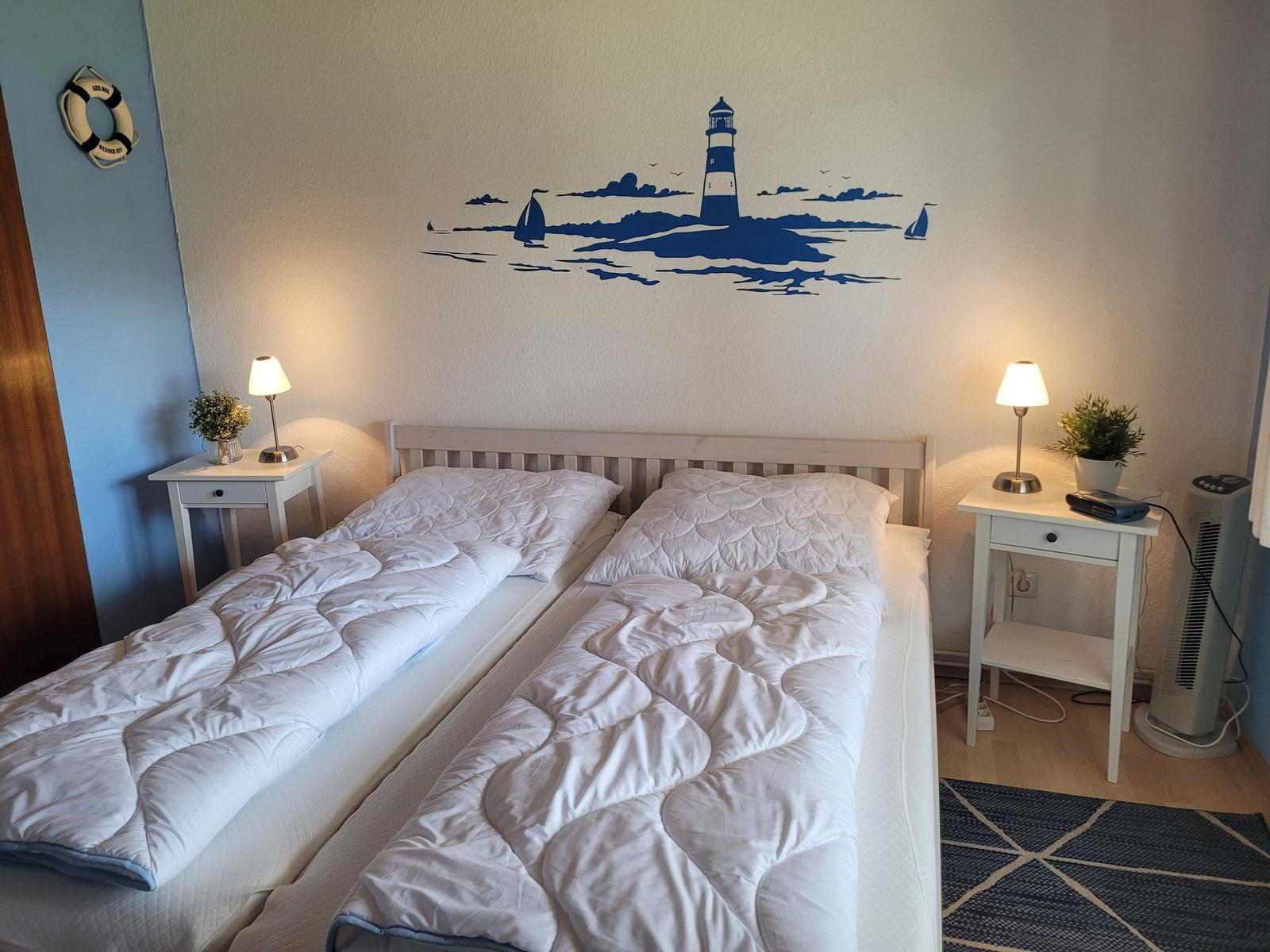 Zwei Betten mit weißen Bettwäsche, Wanddekoration mit Leuchtturm, Nachttische mit Lampen.