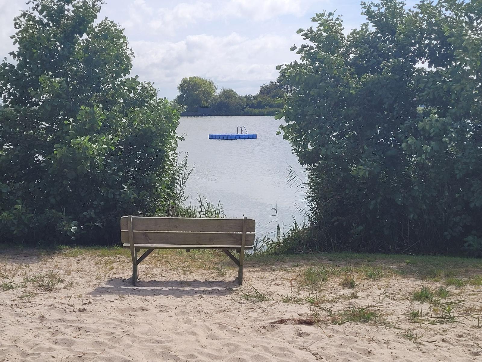 Holzbank am Ufer mit Blick auf einen See und eine schwimmende Plattform.