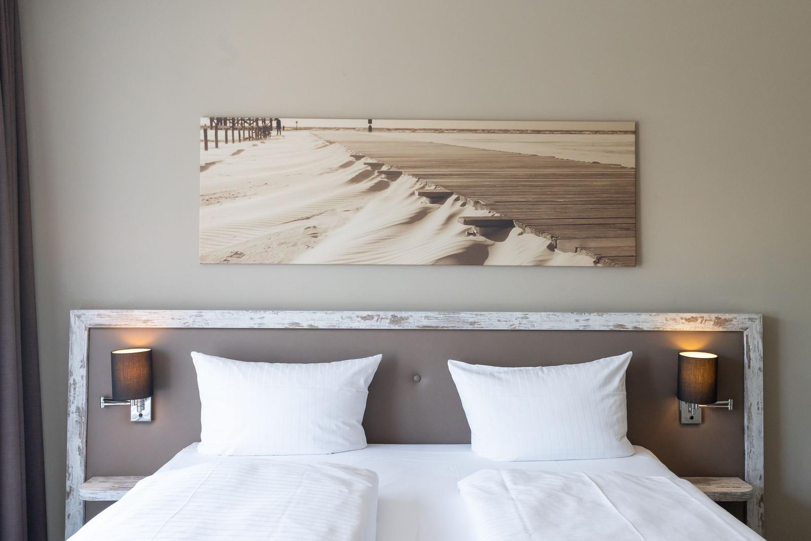 Doppelbett mit weißen Kissen und Kopfteil. Wandbild zeigt Strand mit Holzweg.