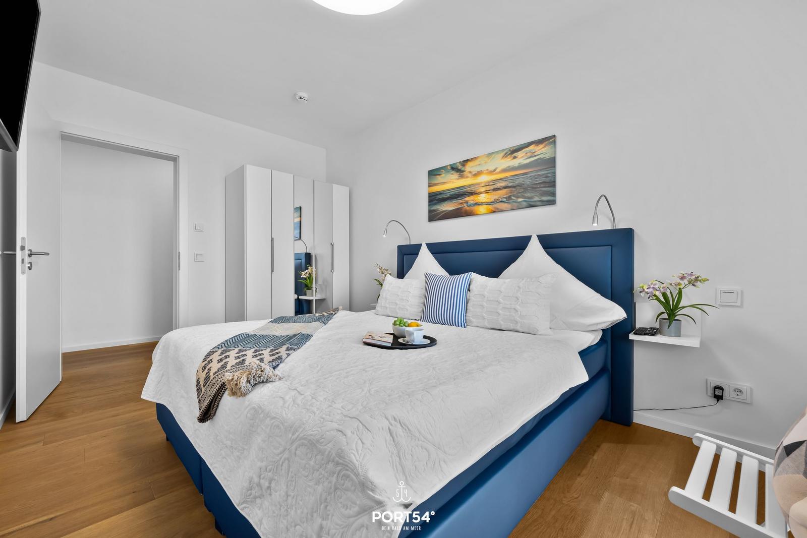 Schlafzimmer mit blauem Bett, weißen Wänden und Holzboden. An der Wand hängt ein Bild.