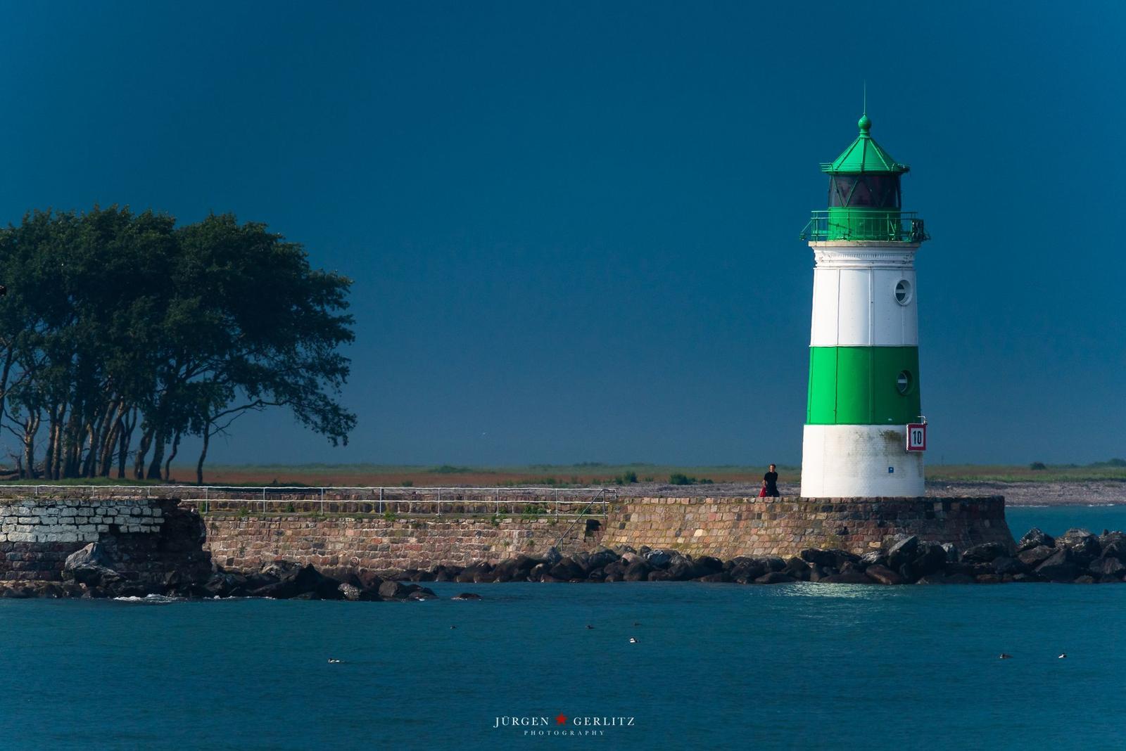 Weiß-grüner Leuchtturm steht auf Steinhafeneinfahrt. Hinter ihm Bäume und blauer Himmel.