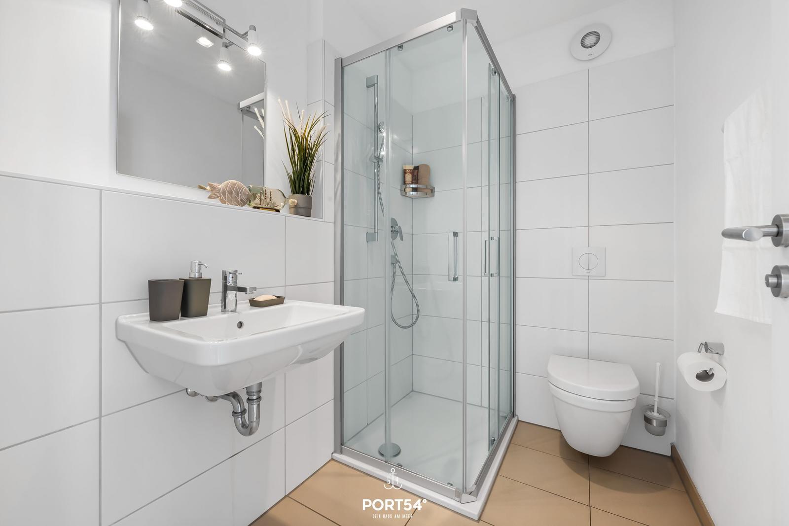 Badezimmer mit Dusche, Waschbecken und WC. Weiße Fliesen und Holzboden.